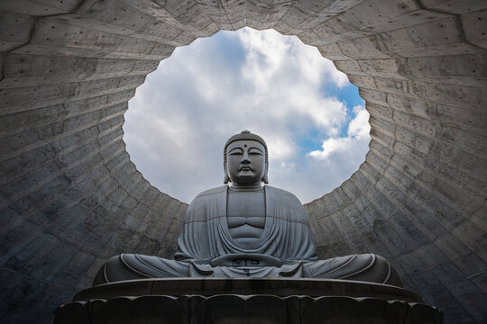 Epic low angle view of the giant Atama Daibutsu (Hill of the Buddha) statue framed by a minimalist concrete dome with a blue sky in the center in Sapporo, Japan
