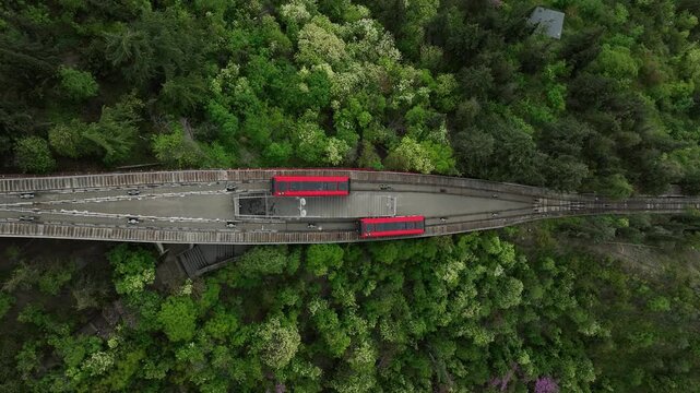 High angle aerial view of Tbilisi funicular tram cars passing each other on Mtatsminda hill, red railway cabins surrounded by green forest and city residential area, Georgia cityscape.