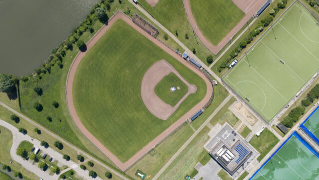 Aerial view of a baseball diamond and multiple green sports fields with a parking lot and solar-paneled building in Houten, Utrecht, Netherlands.
