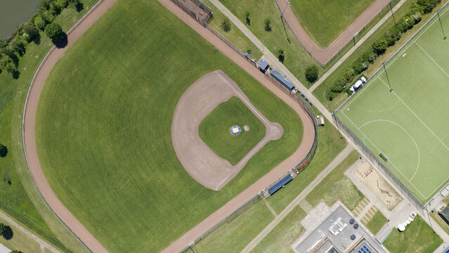 Aerial view of a baseball field with a green outfield and brown dirt infield diamond surrounded by fences and adjacent sports facilities in Houten, Utrecht, Netherlands.