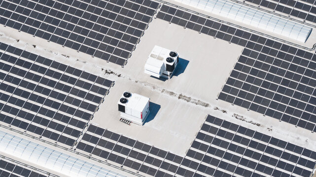Aerial view of a large array of solar panels installed on an industrial rooftop with HVAC units under bright sunlight in Houten, Utrecht, Netherlands.
