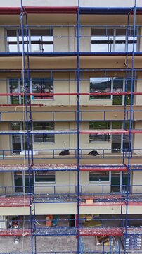 Workers are constructing a multi-storey building using scaffolding. Windows are being installed while construction materials are placed around the site. The setting is urban and busy.