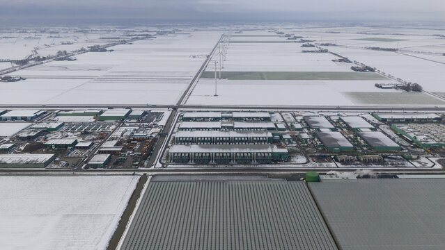 Aerial view of industrial buildings and greenhouses surrounded by snow-covered fields and wind turbines under an overcast sky in Middenmeer, North Holland, Netherlands.