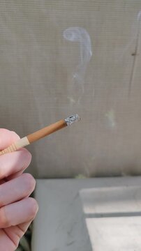 Closeup of a hand holding a burning cigarette with white smoke rising against a blurred background. Bad habit, addiction, and health risks concept for medical awareness or lifestyle contexts.