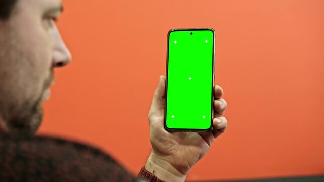 Hand of young man holding smartphone green screen in business office work. Mobile phone vertical mockup. Male person using phone with blank green background display. Chroma key smart phone. Close up.