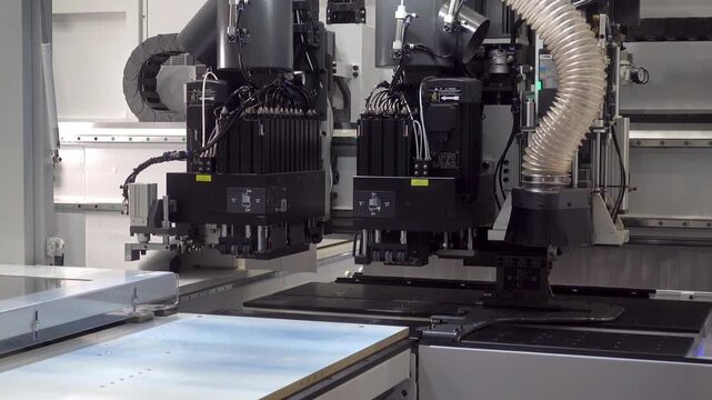 Automated CNC woodworking machine moves a flat wooden panel across the bed while multiple cutting heads and a dust extraction hose operate in a factory setting