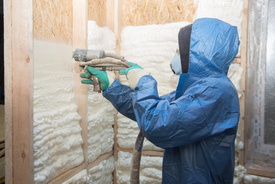 A construction worker in overalls applies insulation foam to a wooden wall of a house, house construction and insulation