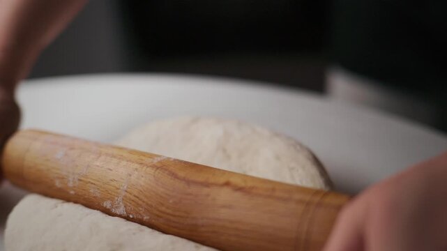 Hands roll smooth dough with wooden roller. Focused effort shown in close-up. Perfect for baking tutorials, recipe videos, or food-themed projects