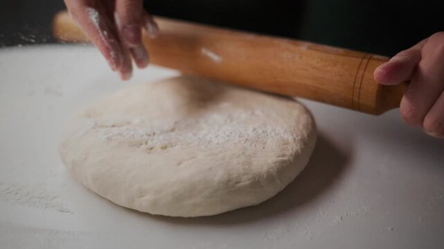 Hands roll dough with wooden pin on floured white surface. Flour dusts as dough flattens. Focus on baking prep. Ideal for cooking, food, or lifestyle videos