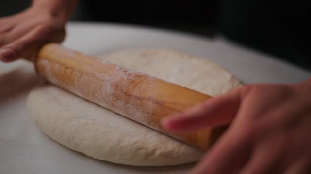 Hands roll dough with wooden roller on white surface. Dough is soft and flour dusted. Action shows careful, patient effort to shape bread base. Suitable for cooking, baking. Ideal for recipe videos