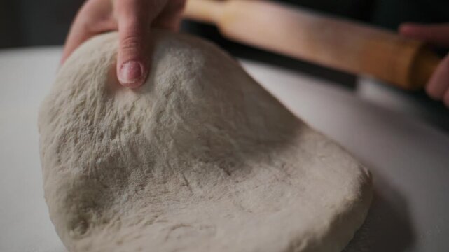 Close-up of hands rolling dough with wooden roller on white surface. Fingers press dough gently, creating soft texture. Dough appears smooth and slightly elastic. Person seems focused and calm