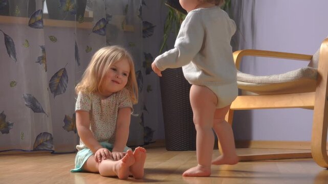 A baby takes first uncertain wobbly steps walking towards their sister. Sweet sibling bond and family moment