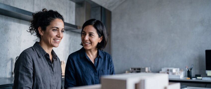 Female architects collaborating in modern studio, professional businesswomen discussing creative architectural design project, smiling women design partners working on construction scale model.