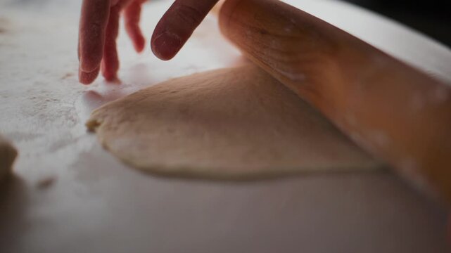 Hand uses a wooden roller to flatten dough on a floured surface. Fingers guide the tool with gentle pressure, ensuring even thickness. Focused, patient work in a warm, homely kitchen