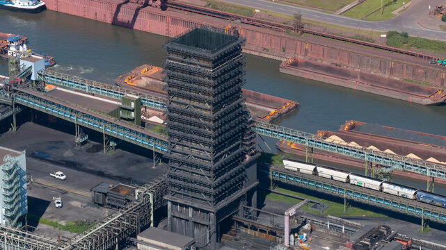 Aerial view of the Kokerei Schwelgern coking plant with its massive tower, conveyor systems, and cargo barges along the industrial waterfront in Duisburg, North Rhine-Westphalia, Germany.