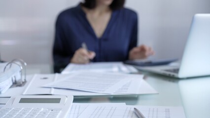 Fototapeta premium Magnifying glass, white calculator, pen and documents are lying on accountant's desk. Businesswoman wearing blue dress is working with documents on the background. Business and audit concept