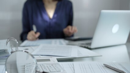 Fototapeta premium Magnifying glass, white calculator, pen and documents are lying on accountant's desk. Businesswoman wearing blue dress is working with documents on the background. Business and audit concept