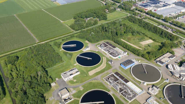 Aerial view of the Rwzi Weurt wastewater treatment plant with circular sedimentation tanks and industrial facilities surrounded by green fields in Weurt, Gelderland, Netherlands.