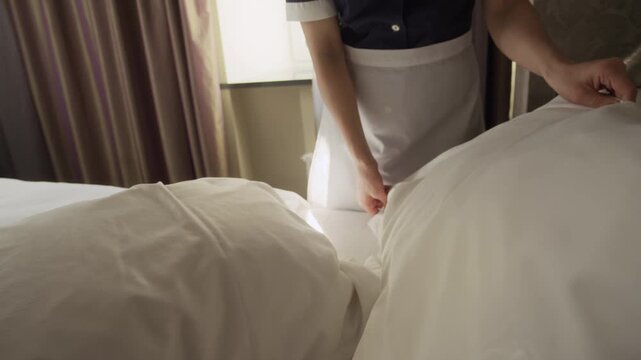 Cropped shot of female housekeeper making bed and fluffing pillows in hotel room. This footage is ideal for hospitality industry marketing, hotel promotional videos, and cleaning service advertisement