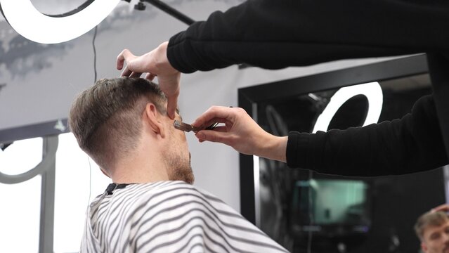 Skilled barber performing precise hair fade using straight razor, working on client's hair in contemporary grooming salon with professional tools