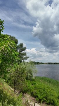 Scenic view of a calm lake with green trees on the far shore and dark grass in the foreground. Beautiful dramatic cloudy sky reflecting in the peaceful water during a summer day outdoors.