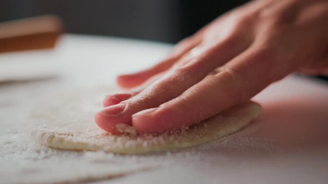 Hand gently presses dough on floured surface. Fingers spread flour as dough flattens. Close-up shot captures texture and motion. Ideal for cooking, baking, or food preparation content