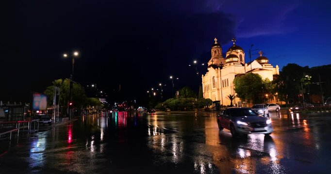 Night video of the illuminated Dormition of the Mother of God Cathedral in Varna city center, Bulgaria during a thunderstorm. Passing cars with headlights on wet asphalt.