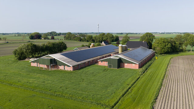 Aerial view of modern farm buildings with solar panels on roofs surrounded by lush green agricultural fields and trees under a clear sky in Giethoorn, Overijssel, Netherlands.