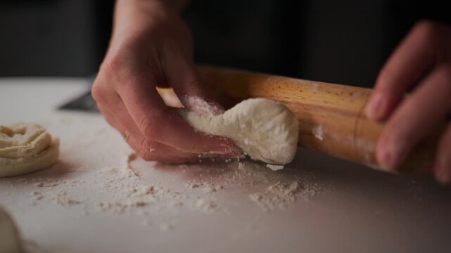 Hands roll dough with wooden roller on floured surface. Fingers guide dough as it flattens. Scene captures cozy, focused kitchen moment. Perfect for recipe videos, cooking tutorials, or bakery content