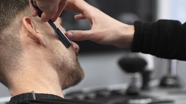 Professional barber carefully shaving client's beard, gripping straight razor with expertise while working in classic barbershop setting