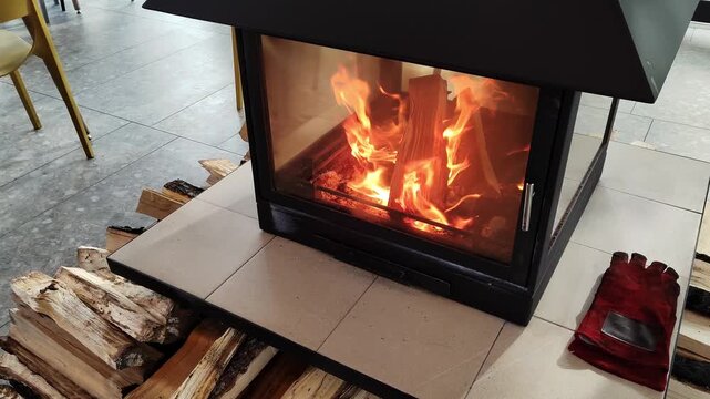 Firewood burns inside a glass fireplace as stacked logs and red gloves rest beside the tiled hearth. The image reflects home heating, winter comfort, energy use, safety, and stress relief.