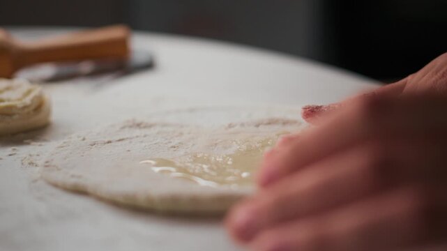 Close-up of hands gently pressing dough on floured surface. Fingers spread dough evenly. Soft focus highlights texture and motion. Ideal for cooking, baking, or food preparation content