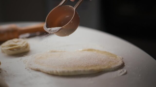 Hand pours flour over a flat dough disk. Dusty white powder settles slowly on the surface. Wooden spoon rests nearby, ready for use. The scene focuses on the texture and motion of flour falling