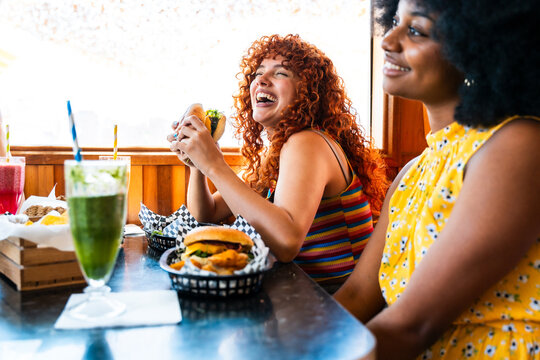 Young multiethnic women laughing enjoying burgers and drinks - Happy multiracial students meeting in a fast food restaurant to have fun together and celebrate