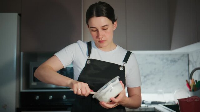 Young woman mixes batter in kitchen. She wears black apron over white shirt. Focus is on hands stirring bowl. Kitchen has modern appliances and marble backsplash