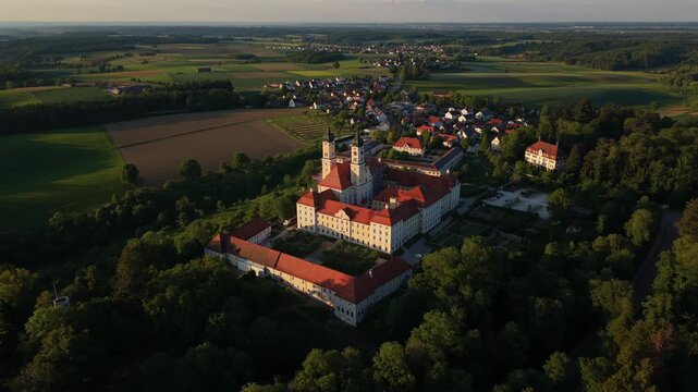 Drone view of Premonstratensian monastery Kloster Roggenburg in Oberschwaben, Germany, baroque architecture, courtyard, farmland and rural landscape, filmed in May with long shadows and soft light