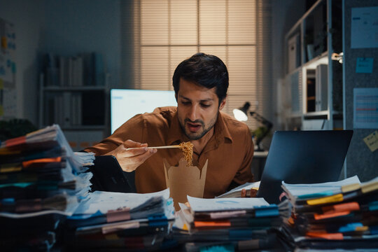 Young Latin businessman eating takeout instant noodles and reviewing document on laptop at desk in dark night office. Overtime working late, overworked deadline, fast food, corporate problem concept.