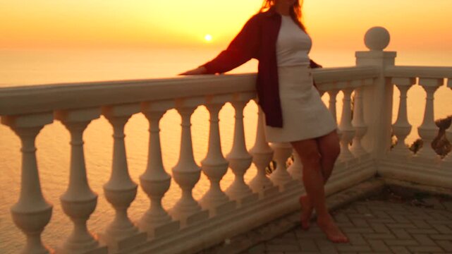 Woman sunset terrace enjoying a breathtaking orange golden hour over the calm ocean from a beautiful classical balcony with balustrade railings