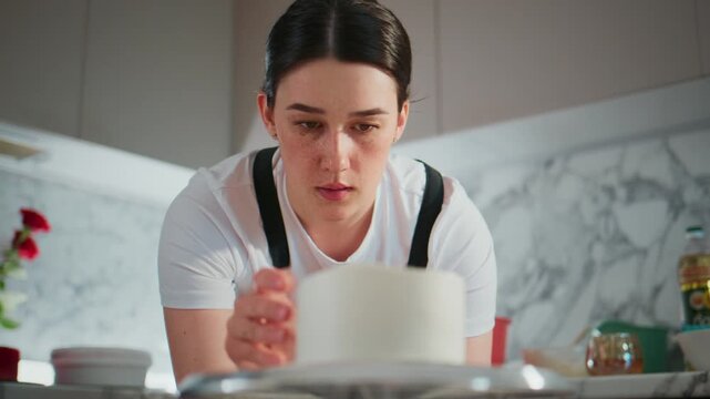 Young woman carefully decorates white cake in modern kitchen. She focuses intensely while holding small tool. Marble backsplash and warm lighting enhance cozy scene. Ideal for baking, celebration