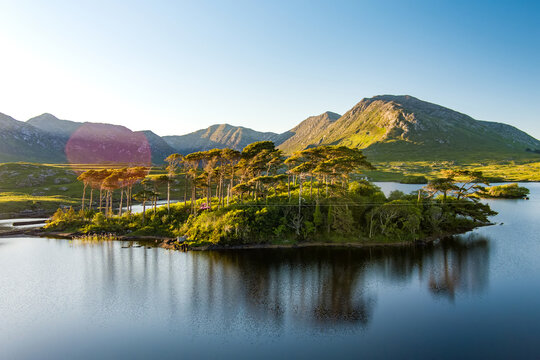Aerial view of Twelve Pines Island resting in still water on Derryclare Lough beneath the Connemara mountains, County Galway. A landscape of an iconic scenic spot in western Ireland.