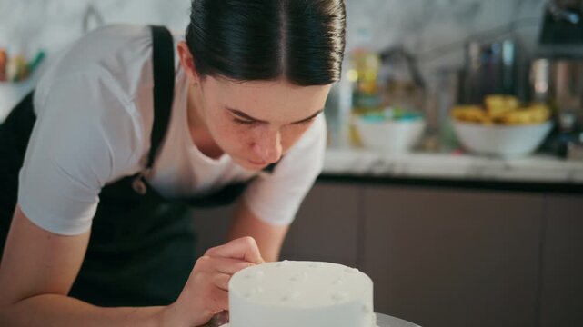 Young woman focuses intently while decorating a white cake. She leans close, showing concentration and care in her craft. Kitchen background includes bowls of fruit and baking tools