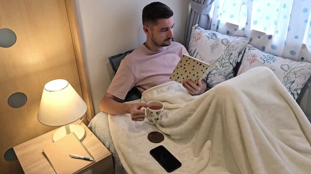 Young man reading book and drinking tea while relaxing in bed under blanket in bedroom. Cozy home leisure footage featuring evening reading routine, comfort and indoor lifestyle