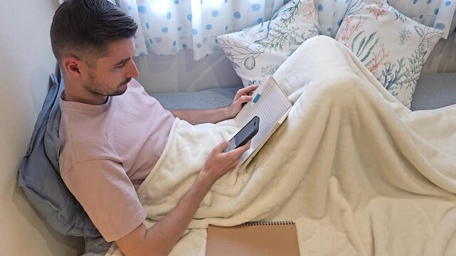 Young man lying in bed under blanket and endlessly scrolling smartphone while holding notebook. Home footage illustrating excessive phone use, screen addiction, doomscrolling, digital distraction