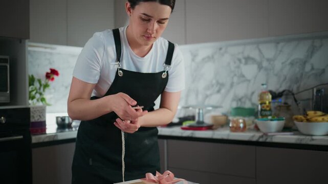 Young woman cuts meat in modern kitchen. She wears black apron and focuses on task. Kitchen features marble backsplash and stainless appliances