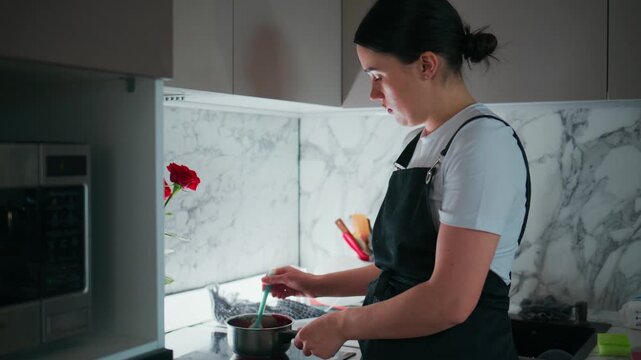 Woman in an apron stirs a pot in a modern kitchen. She focuses intently on her cooking, her expression calm and concentrated. Marble backsplash and kitchen appliances surround her