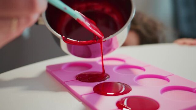 Hand pours vibrant red liquid into a pink silicone mold. Child watches with curiosity from behind. The glossy liquid fills each cavity slowly. Ideal for food crafting, DIY projects