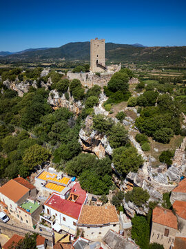 Aerial view of the historic Castello della Fava stone tower standing on a rocky hill surrounded by lush green trees and traditional houses in Posada, Sardinia, Italy.