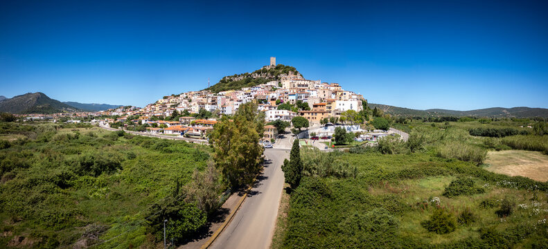 Aerial view of the historic hilltop town of Posada featuring the medieval Castello della Fava tower surrounded by colorful houses and lush green plains in Posada, Sardinia, Italy.