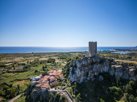 Aerial view of the medieval Castello della Fava tower on a rocky cliff overlooking colorful houses and the blue Mediterranean Sea in Posada, Sardinia, Italy.