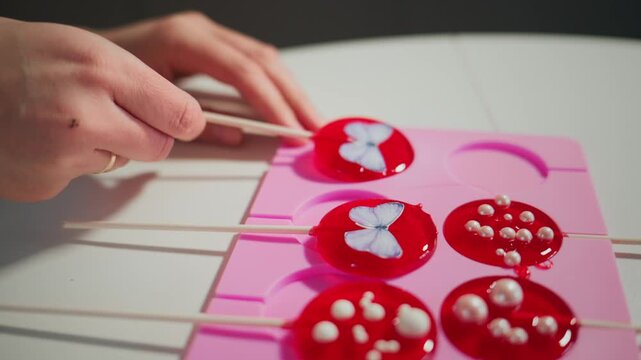 Hands carefully insert sticks into colorful red lollipops. Lollipops feature white butterfly and pearl decorations. Pink mold holds six lollipops. Shot highlights sweet, crafty